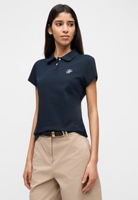 Γυναίκα που στέκεται φορώντας μπλε ναυτικό πόλο πουκάμισο με λογότυπο και μπεζ παντελόνι, κοιτάζοντας ελαφρώς προς τα αριστερά της, μπροστά σε απλό φωτεινό φόντο.