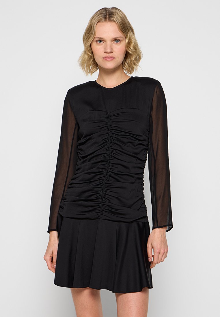 Mujer vistiendo un vestido negro con fruncidos y mangas largas transparentes, de pie contra un fondo claro y liso.