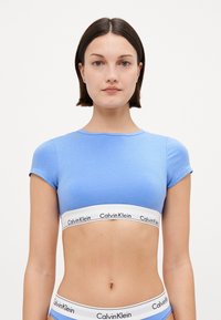 Šviesiai mėlynas prigludęs trumpas viršus su trumpomis rankovėmis, derinamas su baltu elastingu diržu, kuriame išspausdintas juodas tekstas "Calvin Klein". Sklandžios tekstūros, laisvalaikio dizainas.