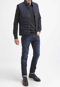 Granatowa bezrękawnik puchowy na teksturowanym czarnym swetrze, zestawiona z ciemnymi d�żinsami i czarnymi butami, prezentująca luźny krój i swobodny styl.