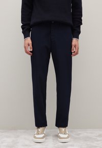 Marineblaue Hosen mit maßgeschneiderter Passform, mit glatter Textur und Knopfverschluss, kombiniert mit beigen und weißen Sneakern.