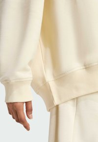 Krémová mikina s rebrovanými manžetami a spodným okrajom, z hladkej textúrnej látky. Ruka umiestnená blízko detailu na spodnom okraji.