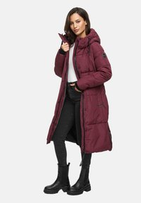 Woman wearing a long maroon padded coat with hood, black jeans, black boots, and a white shirt, standing with one hand holding the collar.