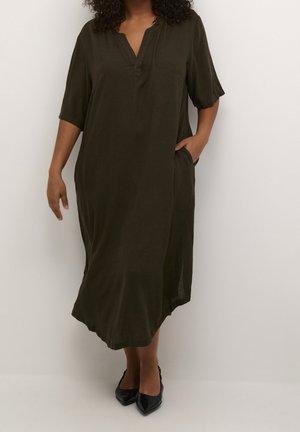 Femme portant une robe midi ample de couleur vert olive foncé avec des manches trois-quarts et des escarpins noirs à bout pointu, debout devant un fond blanc uni.