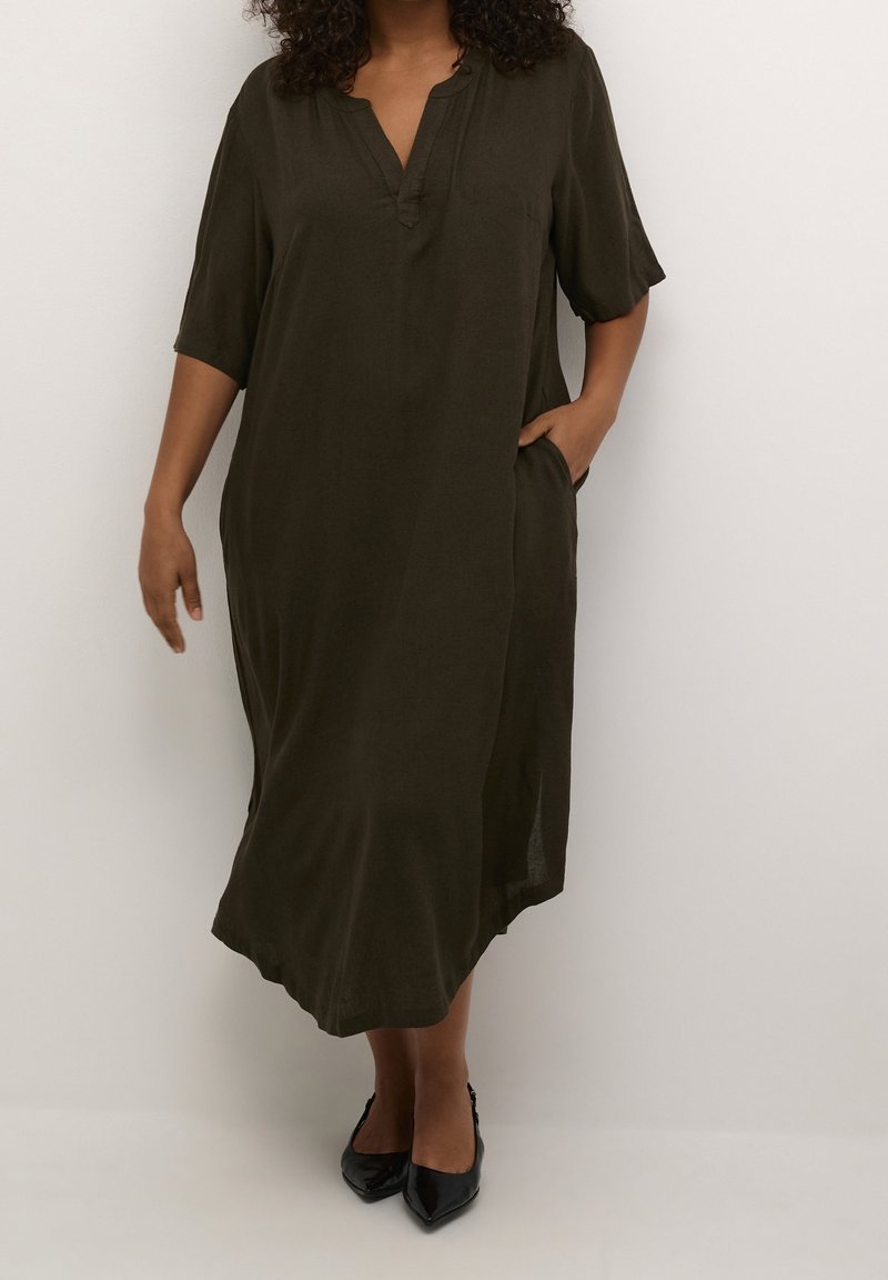 Femme portant une robe midi ample de couleur vert olive foncé avec des manches trois-quarts et des escarpins noirs à bout pointu, debout devant un fond blanc uni.