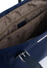 Binnenkant van een marineblauwe stoffen tas met meerdere ritscompartimenten, beige voering met een patroon van het "Hexagona Paris"-logo en stevige hardwareaccenten.