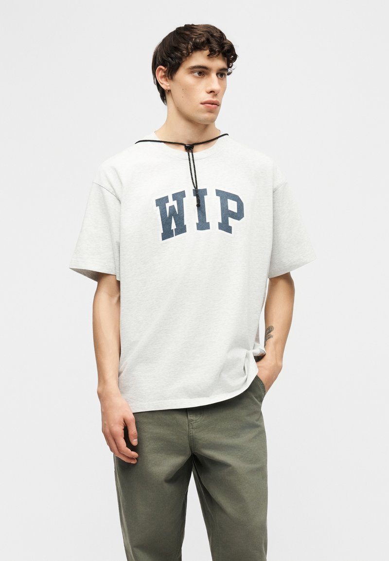 Junger Mann trägt ein weißes, übergroßes T-Shirt mit "WIP"-Druck und olivgrünen Hosen, steht mit einer Hand in der Tasche vor einem schlichten Hintergrund.