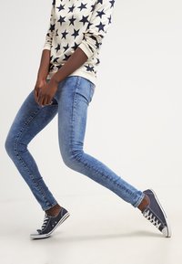 Helle blaue, eng anliegende Jeans mit einem ausgewaschenen Look. Das Modell trägt einen cremefarbenen Pullover mit marineblauen Sternmotiven und marineblauen Sneakers.