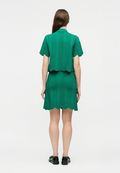 Femme debout de dos, portant un haut vert texturé à manches courtes et une jupe assortie aux bords festonnés, ainsi que des chaussures noires.