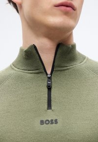 Πράσινο πλεκτό πουλόβερ με ψηλό γιακά και μαύρο φερμουάρ· διαθέτει μαύρο λογότυπο "BOSS" στο στήθος· υφή ύφασματος με ριμπ λεπτομέρειες.
