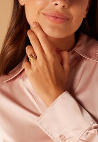 Gouden ring met een ronde, roze steen in een gestructureerde band. De hand van het model steunt op haar gezicht, terwijl ze een lichtroze satijnen blouse draagt.