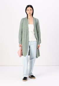 Lång, öppen cardigan i ljusgrön strukturerad stickning, bärs över en vit topp och kombineras med ljusblå jeans och svarta sneakers. Rosa handväska hålls.