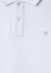 Hvit polo skjorte med teksturert stoff. Har klassisk krage, tre knapper foran og et lys blått brodert emblem på brystet.