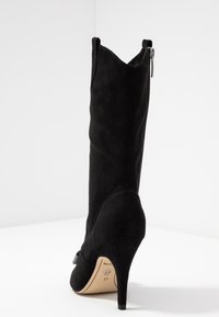 Bottine en daim noir à talon haut avec un embout pointu et une fermeture éclair. Présente une silhouette élégante et un accent de nœud décoratif sur le côté.