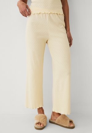 Kremfarvede loungebukser i strik med små hjerteprint, kombineret med matchende top og beige, fluffy åbne tøfler på en stående person.