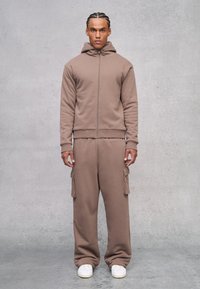 Felpa marrone con cappuccio e zip frontale, abbinata a pantaloni cargo coordinati. Entrambi sono realizzati con un materiale morbido e hanno una vestibilità comoda.