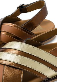 Sandales en cuir marron et or avec larges brides et fermetures réglables à la cheville sur un fond clair.
