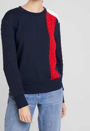 Persoon die een marineblauwe trui draagt met kabelgebreide rode en witte strepen aan de voorkant en op de mouwen, gecombineerd met lichtblauwe spijkerbroek, staand tegen een grijze achtergrond.