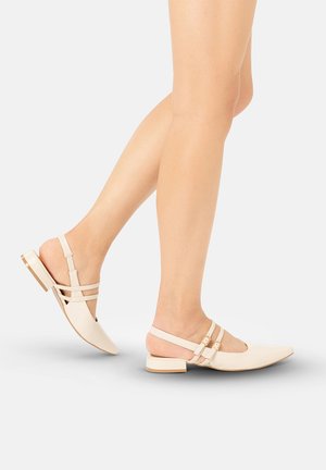 Benen met beige puntschoentjes met slingback, dubbele gespen en lage blokhakken, getoond halverwege een stap tegen een effen achtergrond.