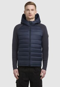 Veste rembourrée bleu marine avec fermeture éclair sur le devant et capuche ; présente une texture matelassée sur le torse et des manches lisses, accentuées par des poignets côtelés.