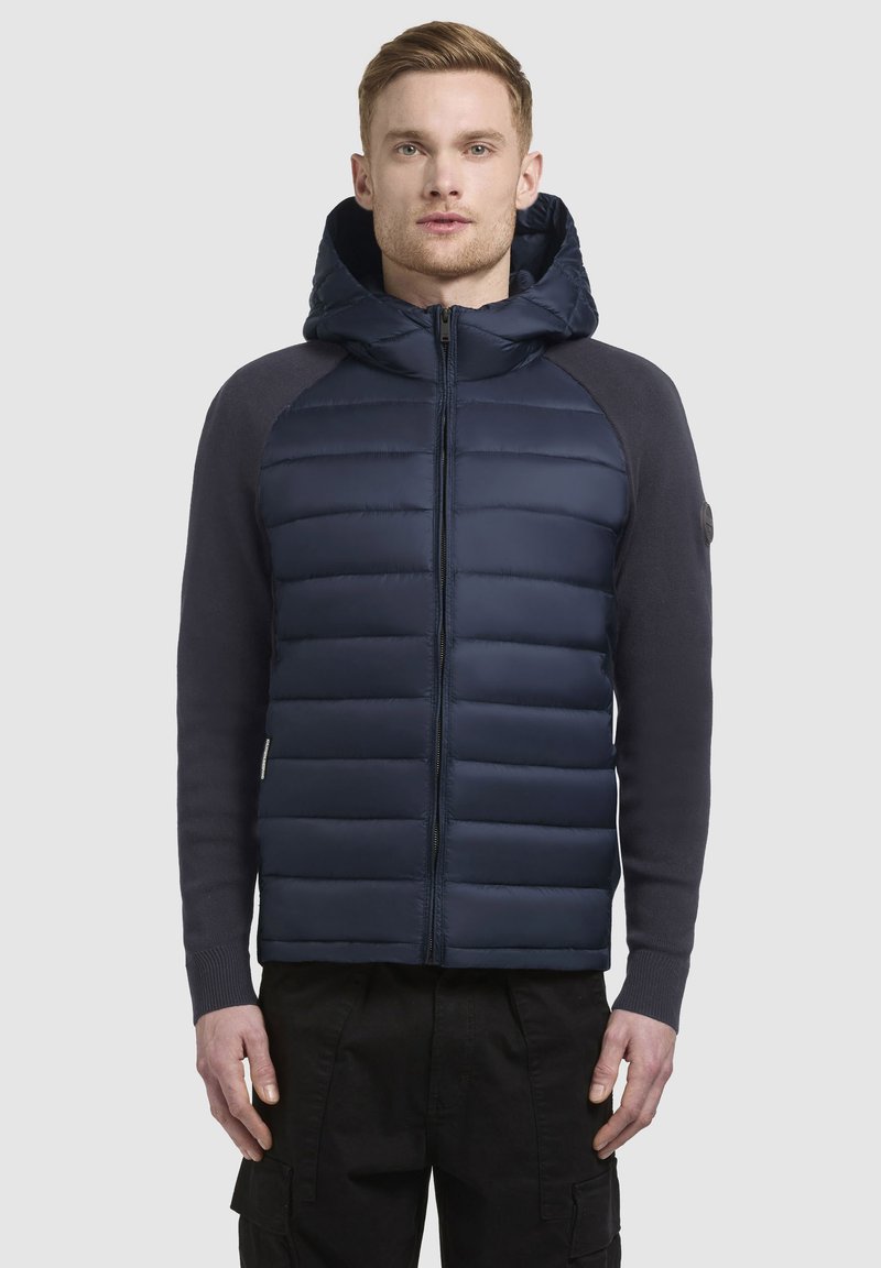Veste rembourrée bleu marine avec fermeture éclair sur le devant et capuche ; présente une texture matelassée sur le torse et des manches lisses, accentuées par des poignets côtelés.