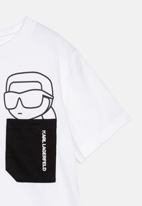 Camiseta de algodón blanca con un bolsillo negro que presenta un diseño de personaje de dibujos animados y el texto "KARL LAGERFELD" en fuente blanca.