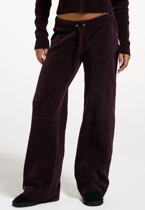 Person trägt dunkelbraune, weit geschnittene Strickhose mit Kordelzug am Bund und passendem kurzen Pullover, steht vor hellem Hintergrund.
