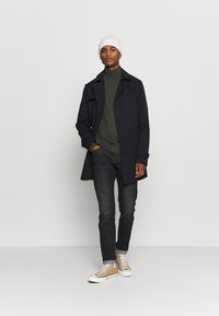 Marineblauwe jas, groene coltrui, donkere jeans, beige hoge sneakers en een lichtroze gebreide beanie. Eenvoudig en modern ontwerp.