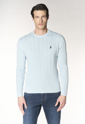 Uomo che indossa un maglione azzurro a trecce e jeans blu, in piedi contro uno sfondo chiaro uniforme, volto rivolto in avanti con espressione neutra.