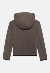 Veste à capuche en tissu brun foncé avec une finition texturée. Présente des coutures décoratives et des finitions noires aux poignets, avec un petit logo noir à l'arrière.