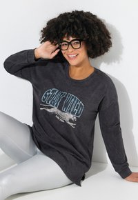 Schwarzes langärmeliges Sweatshirt mit einem grafischen Design eines laufenden Leoparden und Text, kombiniert mit glänzenden silbernen Leggings.
