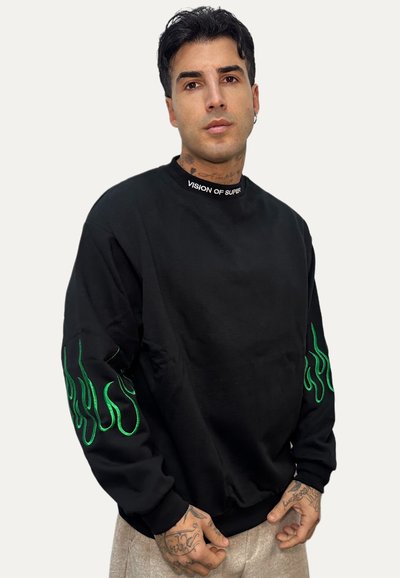 Uomo con tatuaggi che indossa una felpa nera con motivi a fiamma verde sulle maniche e la scritta "VISION OF SUPER" sul colletto.
