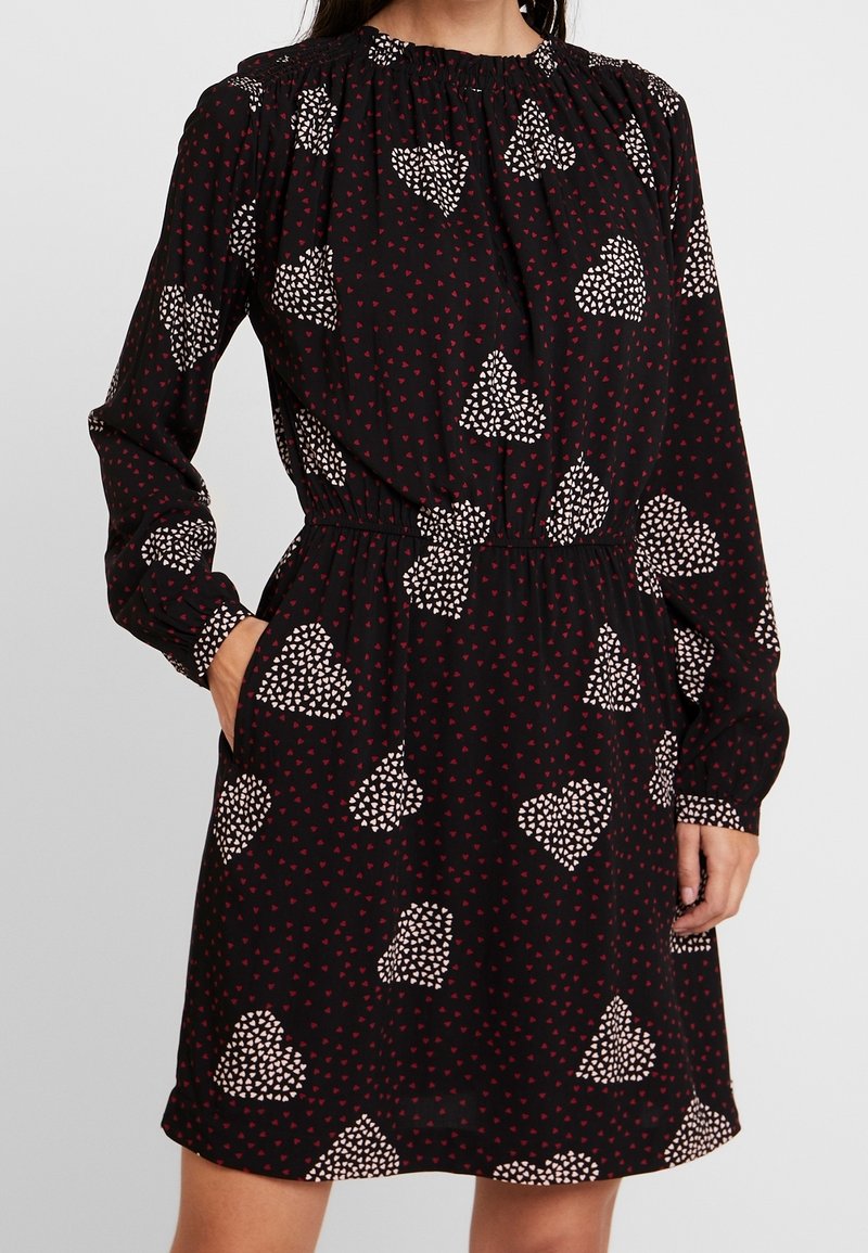 Robe noire à manches longues, col froncé, taille élastique et motif de cœurs rouges avec des formes abstraites blanches. Comprend des poches latérales.