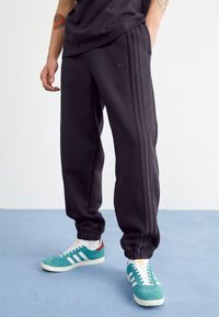 Pantalones deportivos negros con tres franjas laterales, puños ajustados y un pequeño logo bordado. Llevados con zapatillas turquesas que cuentan con franjas blancas.
