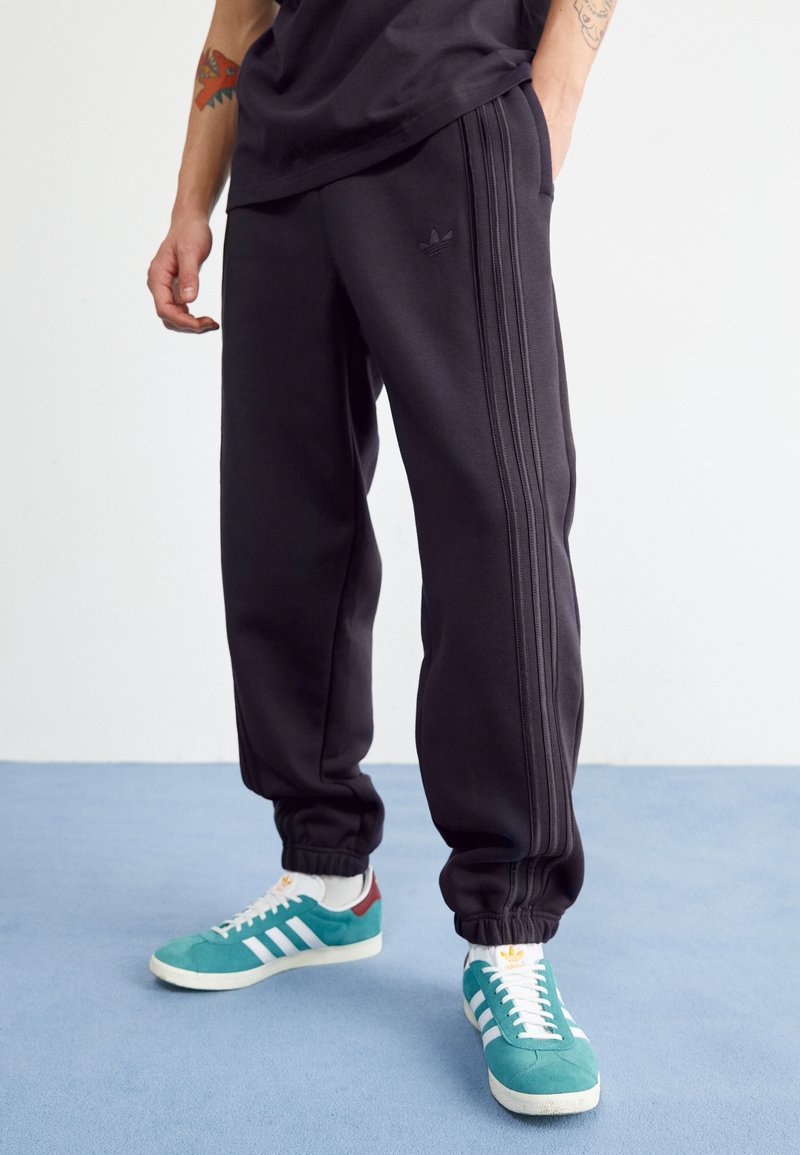 Pantalones deportivos negros con tres franjas laterales, puños ajustados y un pequeño logo bordado. Llevados con zapatillas turquesas que cuentan con franjas blancas.
