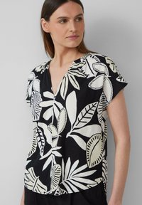 Zwarte blouse met korte mouwen, voorzien van een wit bladerenpatroon, met een V-halslijn en een losse pasvorm. De stof lijkt lichtgewicht en gestructureerd te zijn.