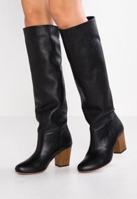 Bottes en cuir noir, montantes jusqu'au genou, avec une surface texturée, un bout arrondi et un talon en bois robuste. Fini lisse avec des plis discrets.