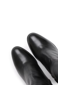 Bottes en cuir noir montant jusqu'au genou, à finition lisse, bout pointu et détails de couture minimalistes, posées sur un fond blanc.