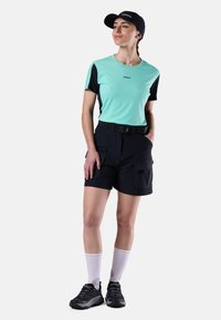 Frau trägt ein mintgrünes und schwarzes kurzärmeliges Hemd, schwarze Shorts mit Gürtel, schwarze Mütze, weiße Socken und schwarze Sportschuhe, steht mit den Händen in den Taschen.
