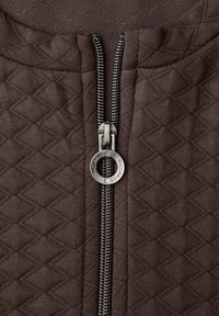 Zbliżenie na brązową pikowaną tkaninę z metalowym suwakiem w kształcie okrągłego pierścienia, wyrytą napis "Street One".