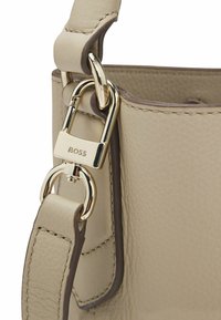 Beige leren handtas met gouden beslag, voorzien van een opvallende sluiting en contrasterende donkerbruine accenten op de riem en randen.