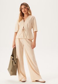 Beige to-delt outfit med en kortærmet top med bindebånd og vide bukser. Modellen holder en olivengrøn taske.