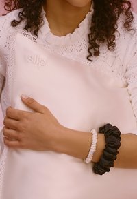 Almohada de seda en suave rosa con un ligero relieve que dice "desliz". Una persona la sostiene, vistiendo una goma de pelo blanca y otra negra en su muñeca.