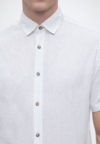 Άνδρας που φοράει λευκό πουκάμισο με κοντά μανίκια, κουμπωτό με ορατά κουμπιά και μυτερό γιακά, κομμένο στο λαιμό και στα χέρια.