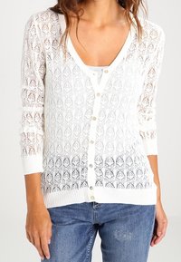Mujer vestida con un cárdigan de botones blanco hecho a crochet sobre una blusa blanca, combinado con jeans desgastados azules, de pie frente a un fondo simple.