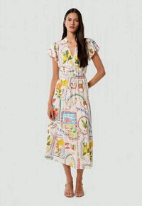 Nainen seisoo päällään vyöllinen, lyhythihainen midi-mekko, jossa on värikäs hedelmien ja kukkien kuvio, yhdistettynä avokärkisiin sandaaleihin.