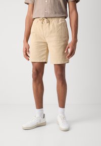 Shorts en velours côtelé jaune clair avec taille élastique et cordon de serrage, finis par un ourlet roulé. Portés avec des baskets blanches et des chaussettes.