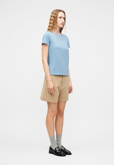 Γυναίκα με κυματιστά ξανθά μαλλιά, φορώντας ανοιχτό μπλε μπλουζάκι, μπεζ σορτς, γκρι κάλτσες και μαύρα μοκασίνια, στέκεται προφίλ σε απλό φόντο.