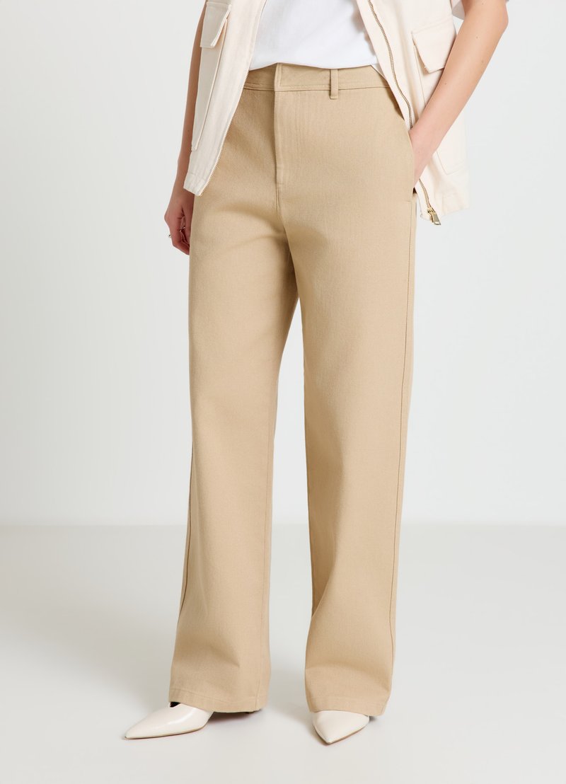 Pantalon beige taille haute à jambes larges avec une texture lisse. Caractérisé par une coupe classique, des passants pour ceinture et un devant épuré. Associé à des chaussures à bouts pointus.