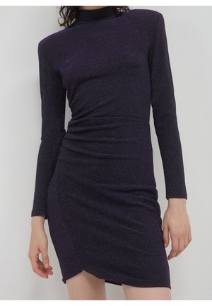 Frau in einem enganliegenden, langärmligen, hochgeschlossenem, dunkel lila glitzernden Kleid mit leichter Raffung und asymmetrischem Saum.